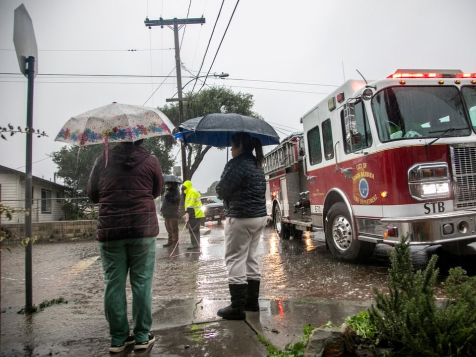 Two women with umbrellas are standing in the rain next to a large fire truck,