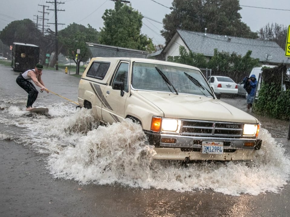 An off-road vehicle pulls a man on a surfboard through a flooded street.