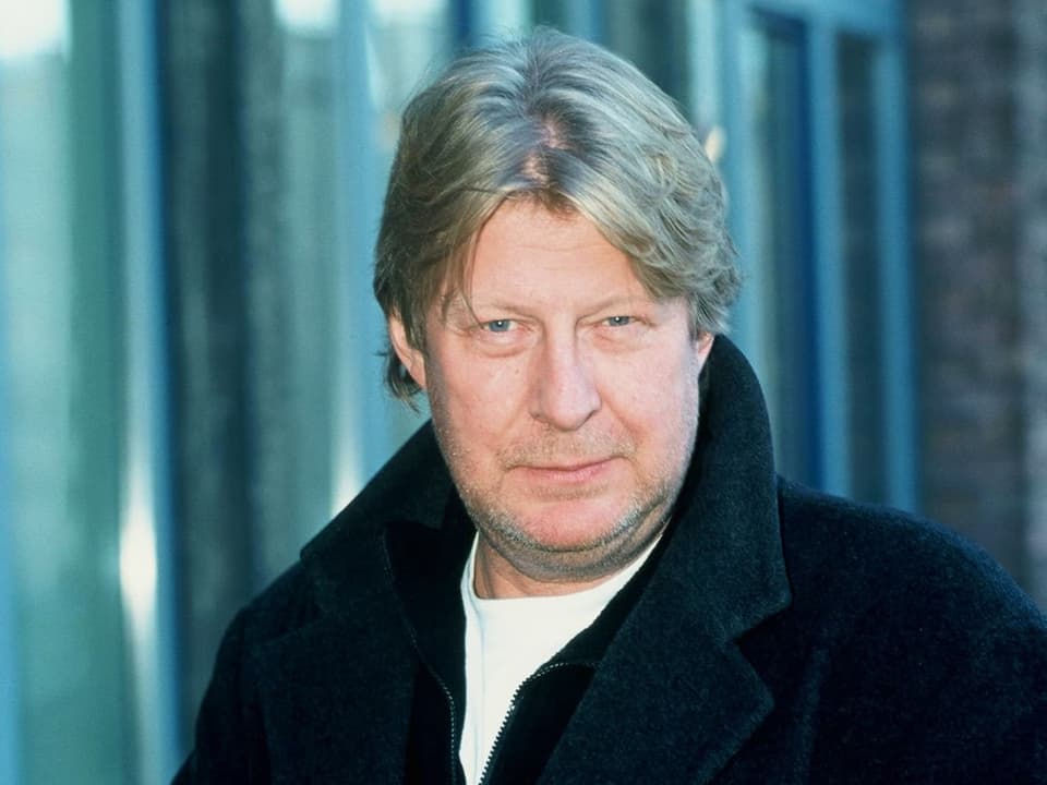 A man in a black coat looks into the camera with slightly narrowed eyes.