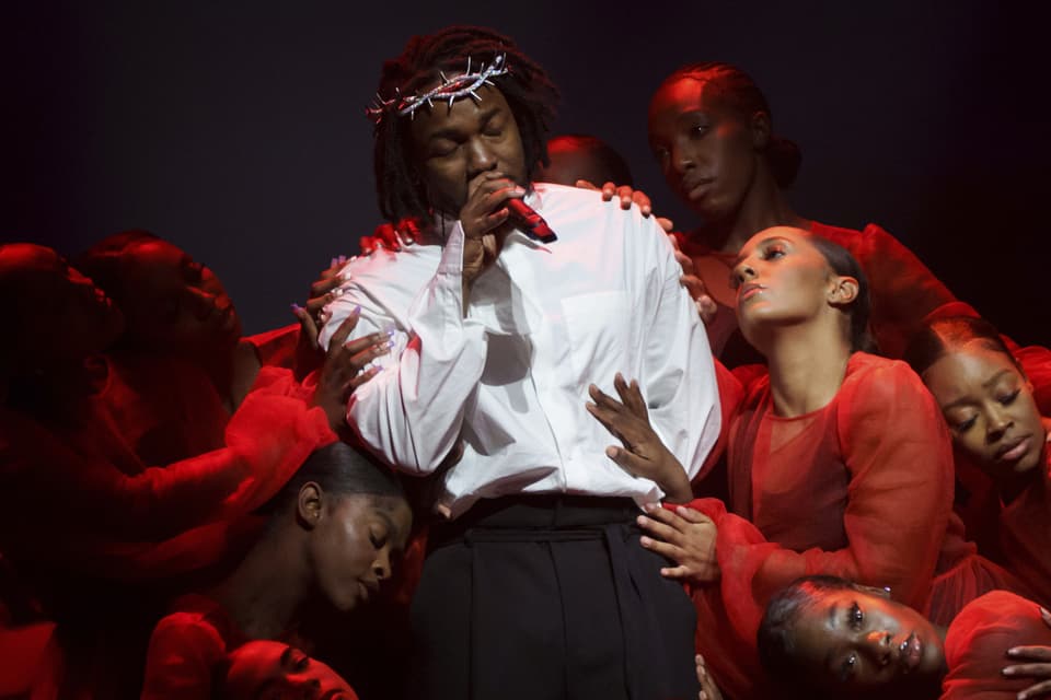 Kendrick Lamar wears a white shirt and crown, surrounded by women dressed in red.