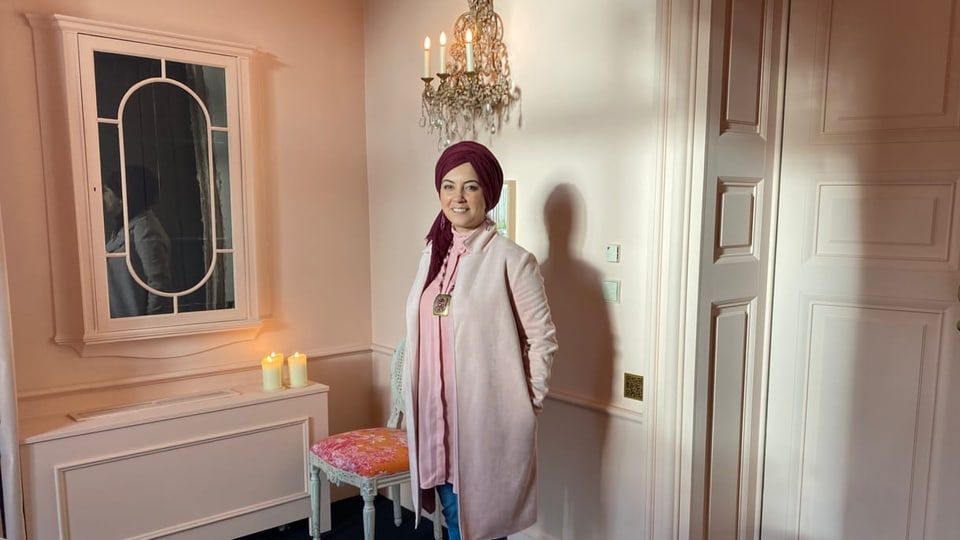 Fatima Zibouh is standing in a room wearing pink dresses.  A burgundy headscarf. 