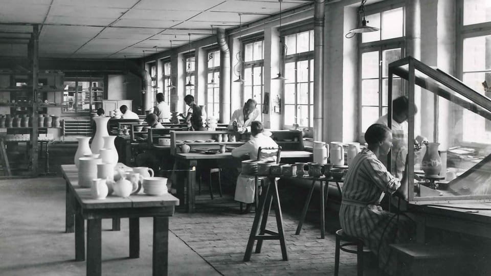 A look into a factory where men and women at workbenches clay and clay jugs.