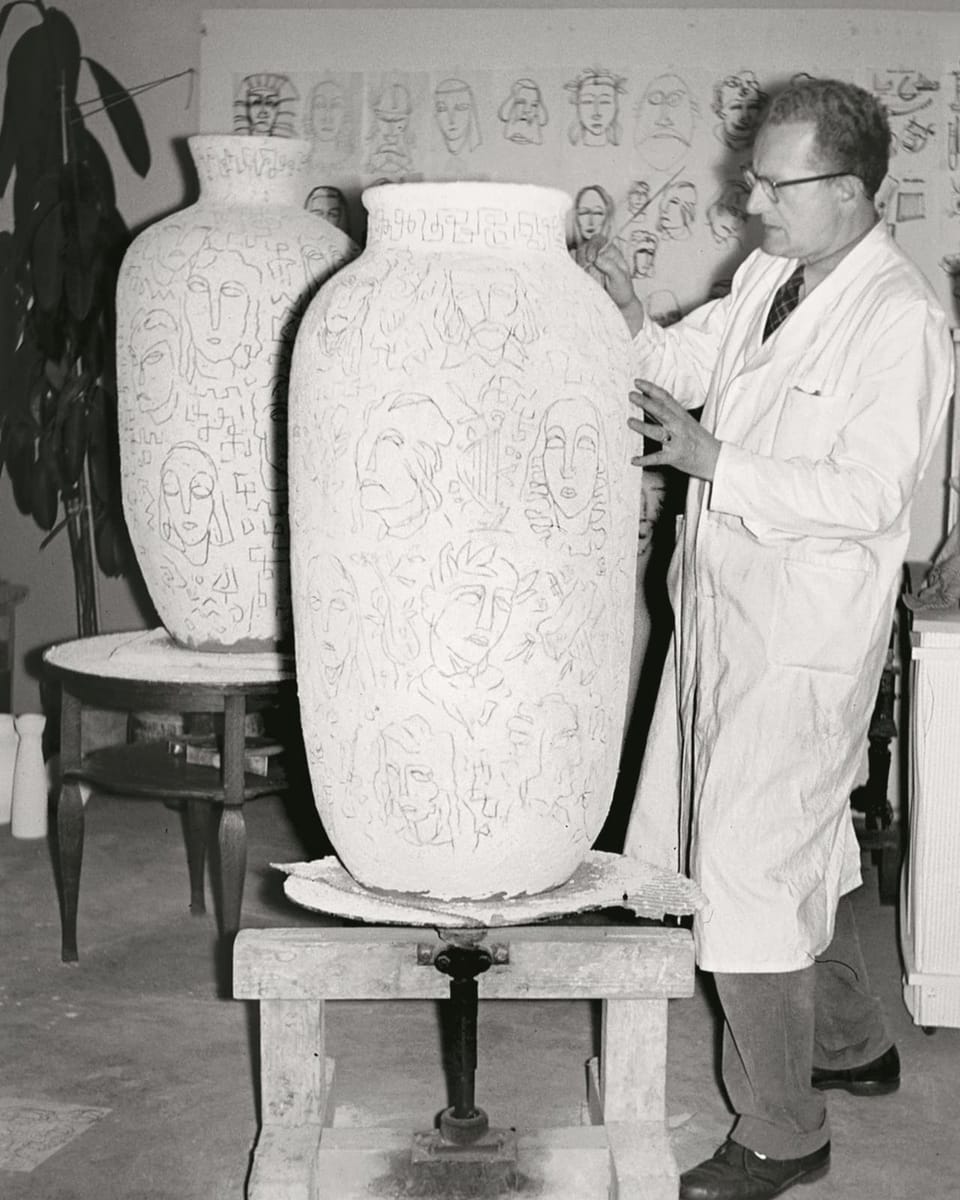 A man dressed in a white overall is working on a waist-high vase.  The motifs show faces. 