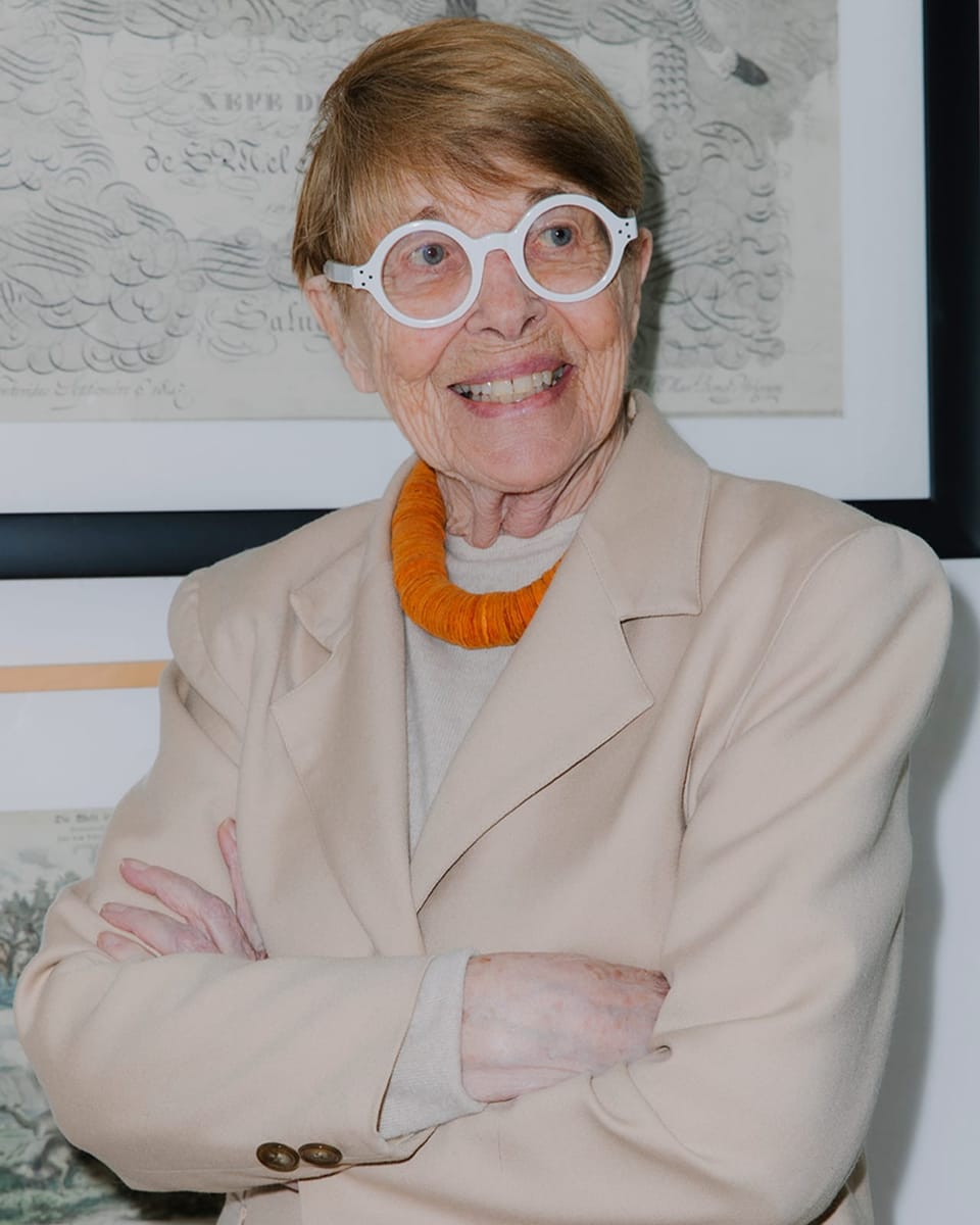 older woman in beige blazer and orange glasses, arms crossed