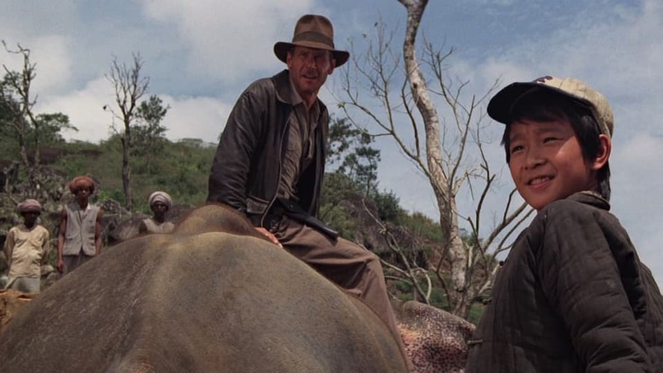 Indiana Jones is seated on the back of an elephant, to his right is a young boy in a baseball cap