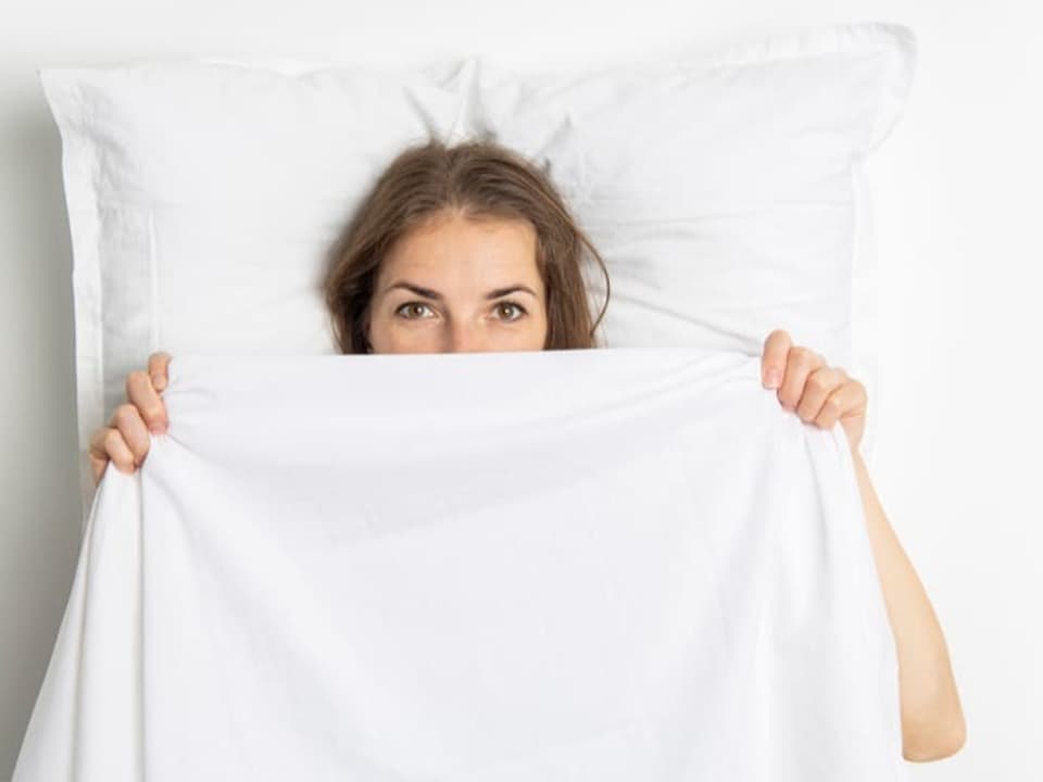 A woman lies in bed and pulls the blanket over her eyes.