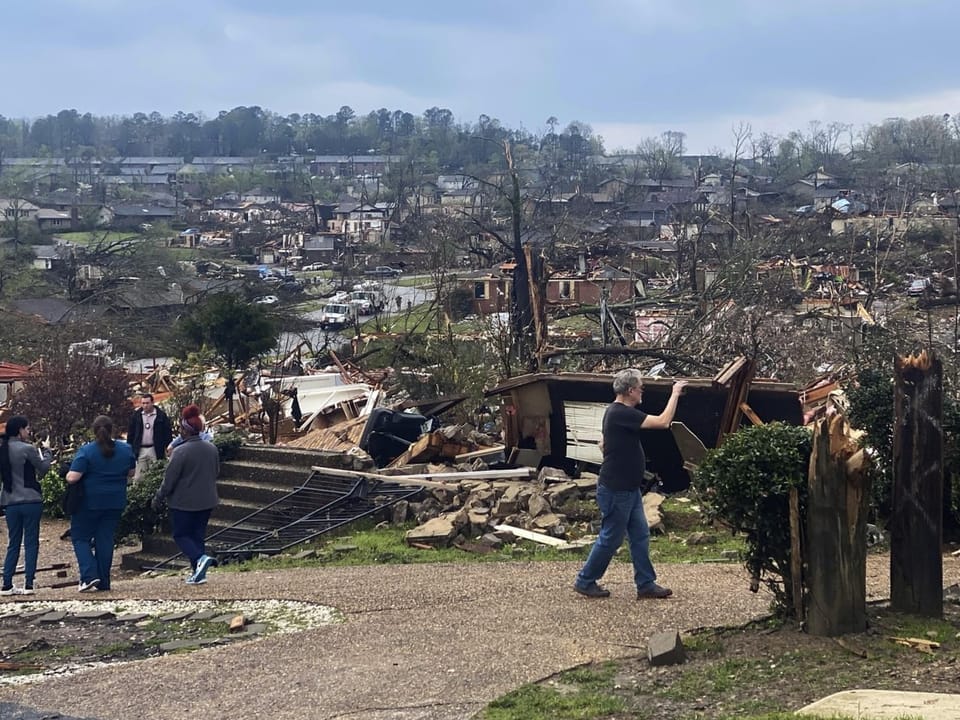 People walk past a collapsed wooden hut.