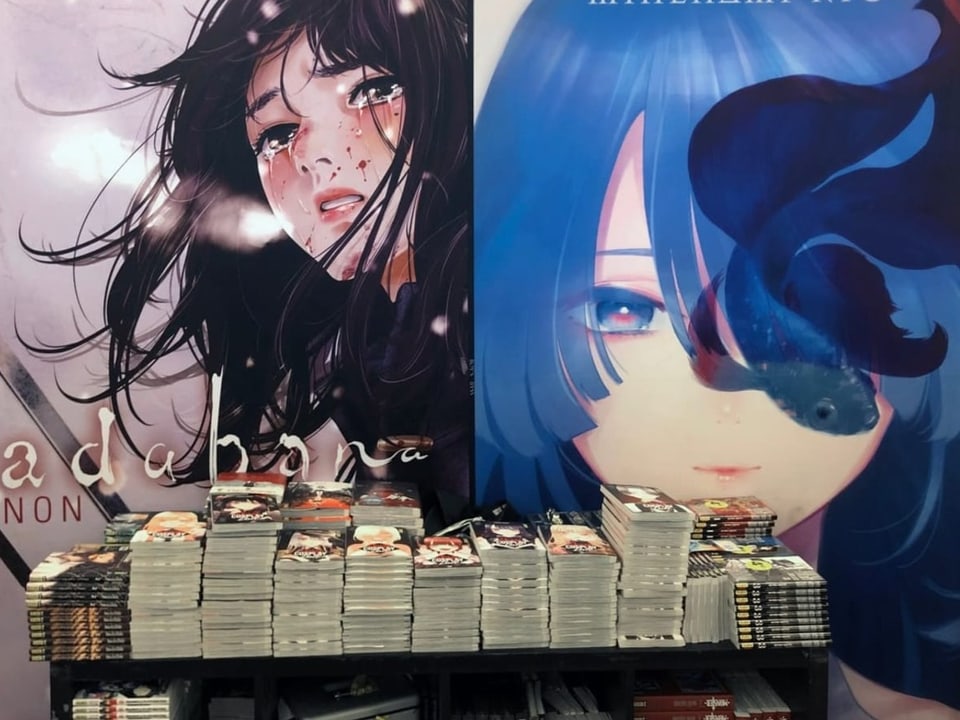 Manga are stacked on a table.  In the background are large advertising posters of manga books.