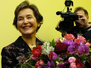 Elisabeth Kopp smiles and carries a bouquet of flowers while a journalist films her.
