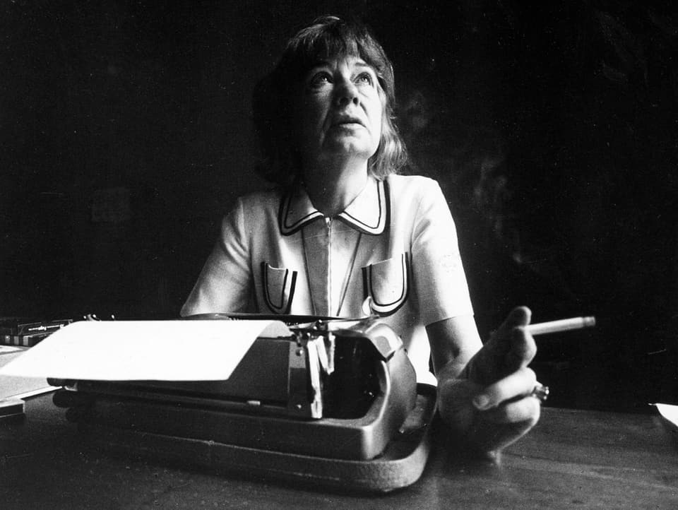 An elderly woman looks up from her typewriter.  She holds a cigarette in her left hand.