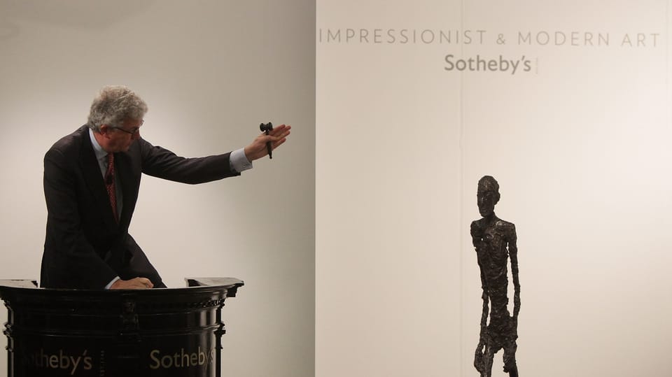 A man in a suit and holding a hammer raises his arm to a metal sculpture on his left.