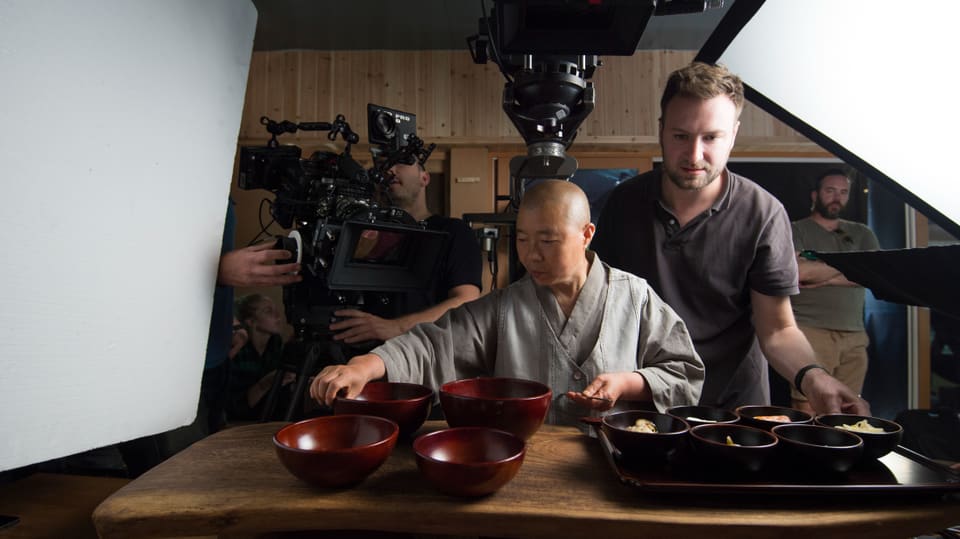 in the middle a nun in a gray robe, bending over red bowls.  Right and left cameramen filming.