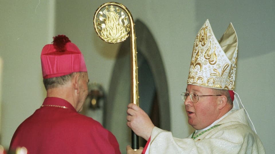 A man in a red velvet robe hands over a shiny gold miter and a curved staff to a bespectacled man.