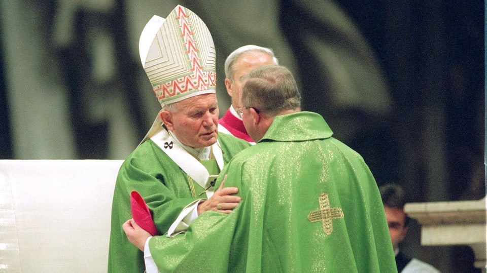 An old man with a miter on his head and a green priest's robe embraces another older man, also in green.