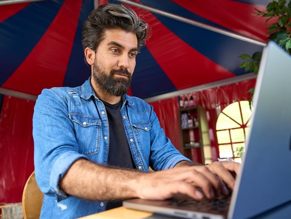 Man sitting in tent at laptop, smiling and typing. 