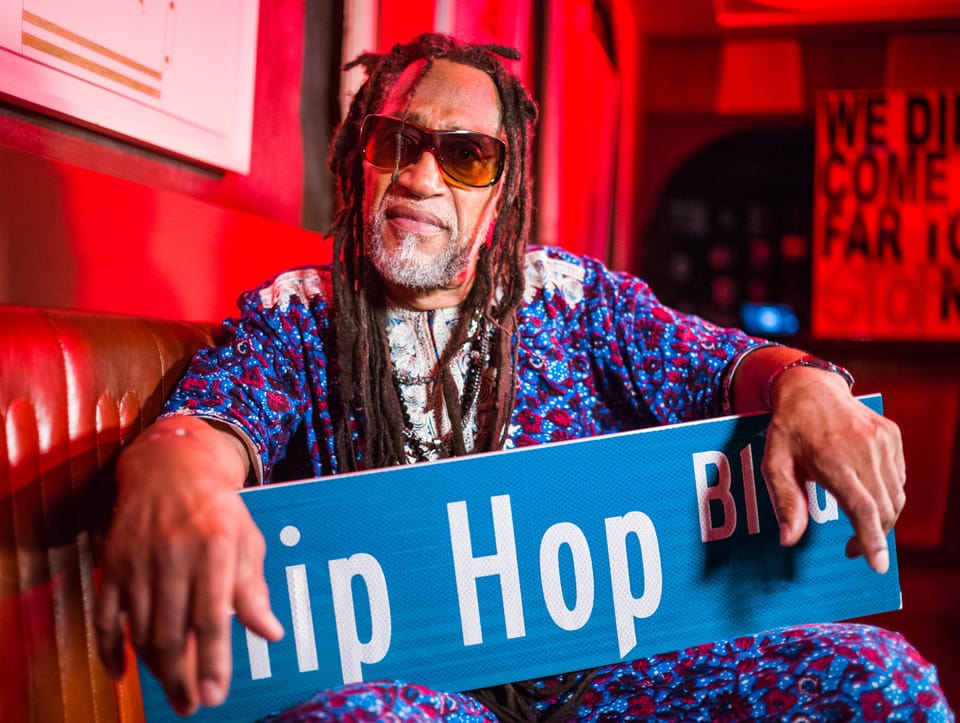 älterer, schwarzer Mann mit Rastas auf roter Couch mit Blau-rotem Hemd, hält blaues Schild: «Hip Hop Bvd», Sonnenbrille.