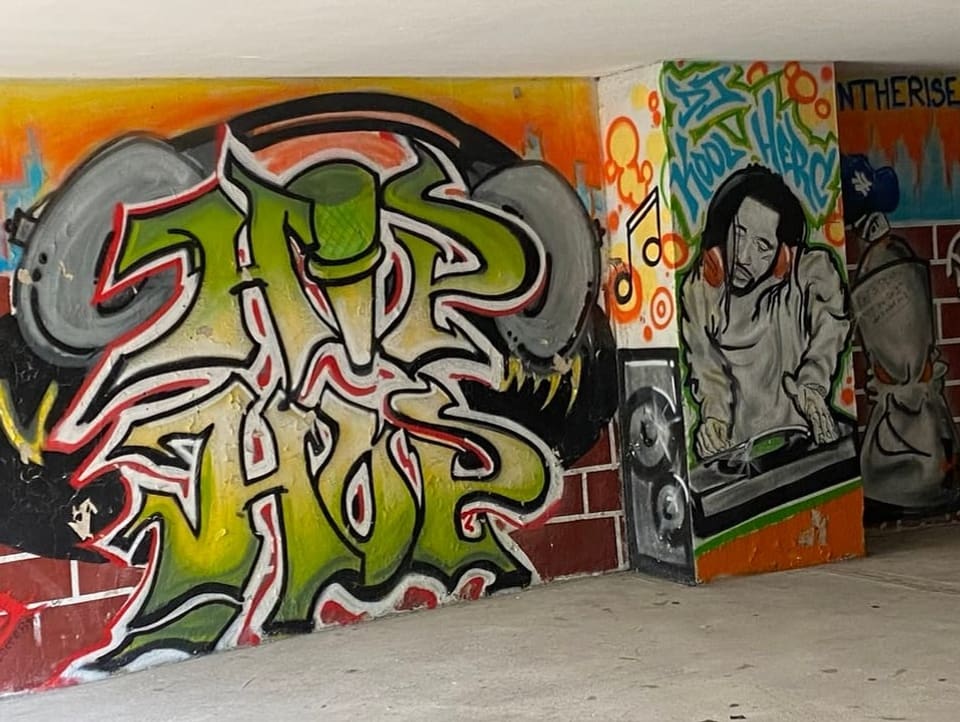 Grünes Graffiti auf der Wand, es steht «Hip Hop», daneben Graffiti eines Typen am DJ-Pult.