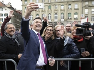 A man takes a selfie with a mobile phone, behind them a lot of people are cheering.