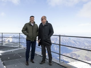 Two men in winter clothes are standing in front of a mountain panorama.