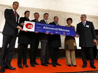 Seven people stand on a red carpet and hold a sign that reads "Lucerne Allmend/Messe".