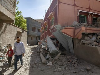 A man walks past a destroyed building with a boy in his hand.