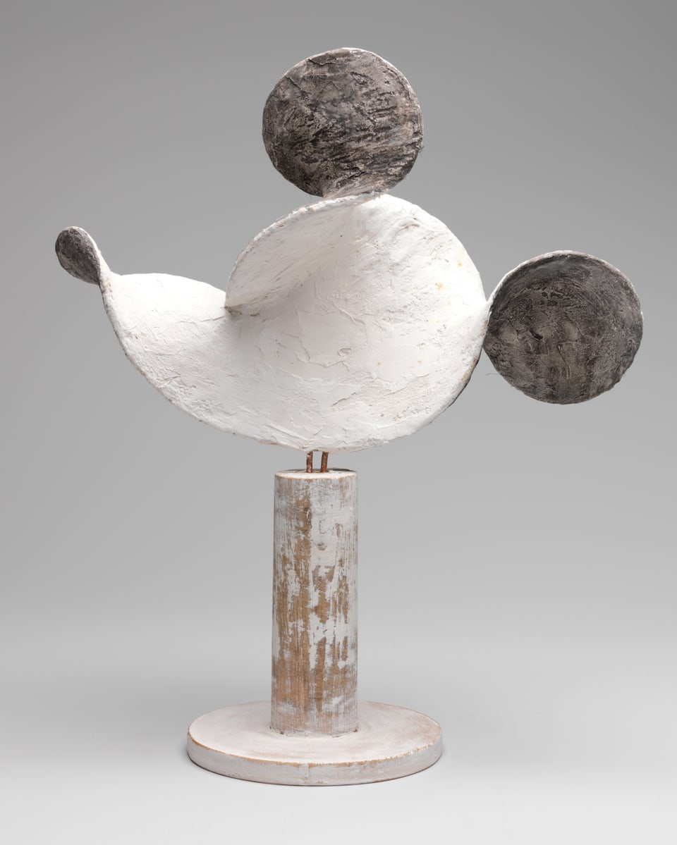 White sculpture with two black circles, looks like a mouse head.  On a wooden, white base.