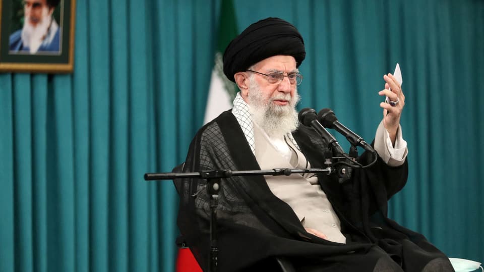 Ali Khamenei vor einem grünen Vorhang. Er sitzt auf einem Stuhl und spricht in ein Mikrofon.