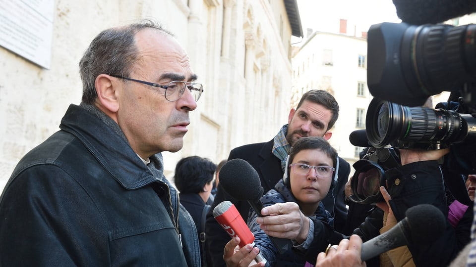 An older man with glasses and a high forehead stands in front of a crowd of journalists with cameras and microphones.