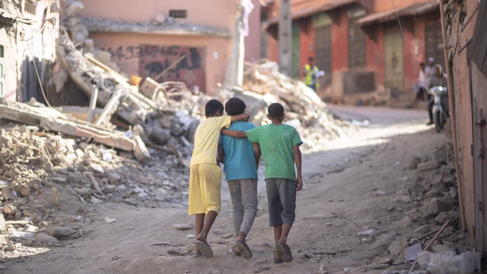 Three boys run arm in arm (photographed from behind) through rubble in a village