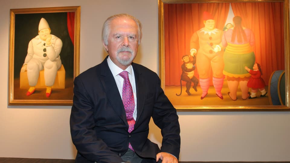 An old man in a suit sits in front of two paintings.