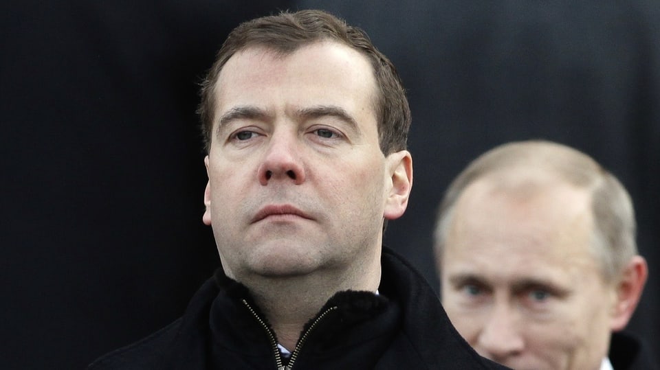 Medvedev looks stern, Putin can be seen behind him.