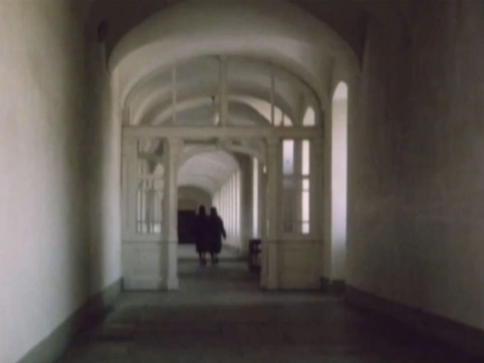 Two people walk down a long hallway.  The image is black and white