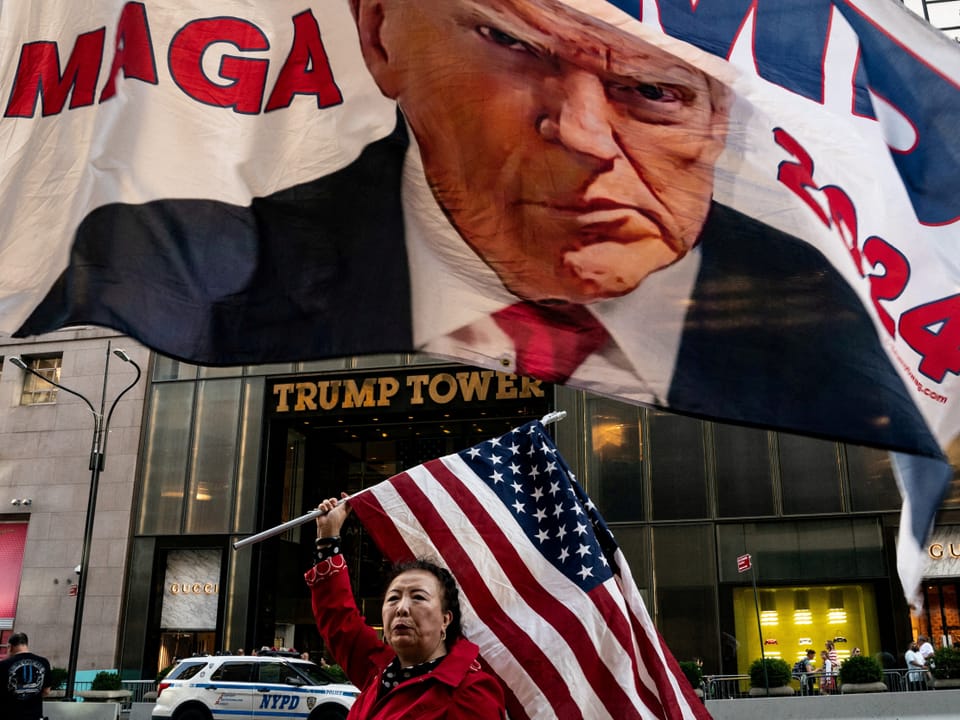 A woman holds the American flag in front of Trump Tower.  A campaign flag with Trump's face flies above her.