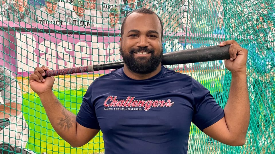Carlos Nepomuceno wears a dark blue Challengers t-shirt.  He holds his baseball bat in both hands.