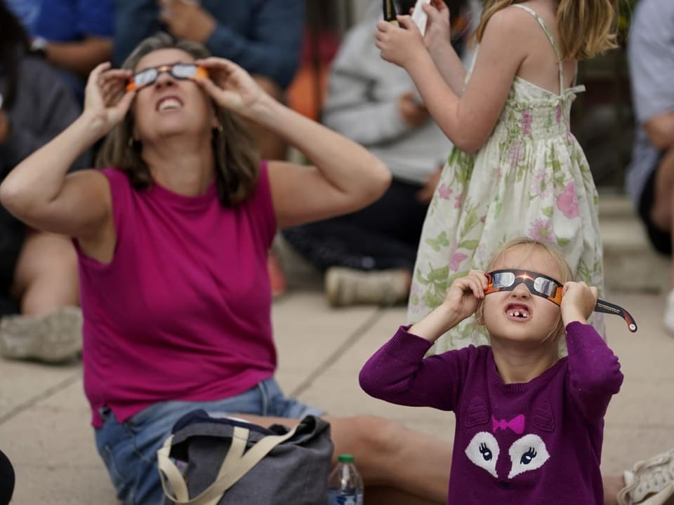 A woman and a child sit next to each other on the floor.  They wear goggles and look at the sky.
