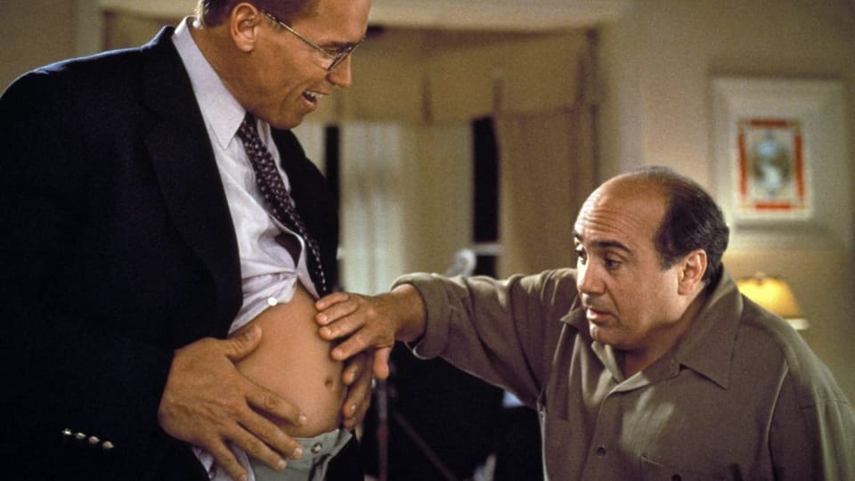 A man in a black suit reveals his baby bump.  A man next to him touches him and is amazed