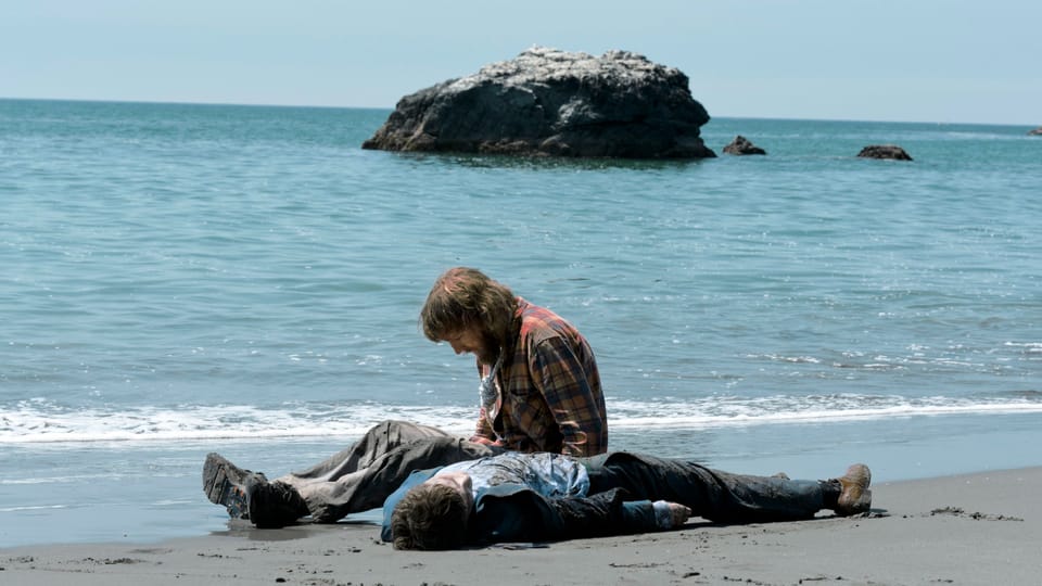 On a beach.  A man lies on the sand, another man sits next to him and looks at the ground
