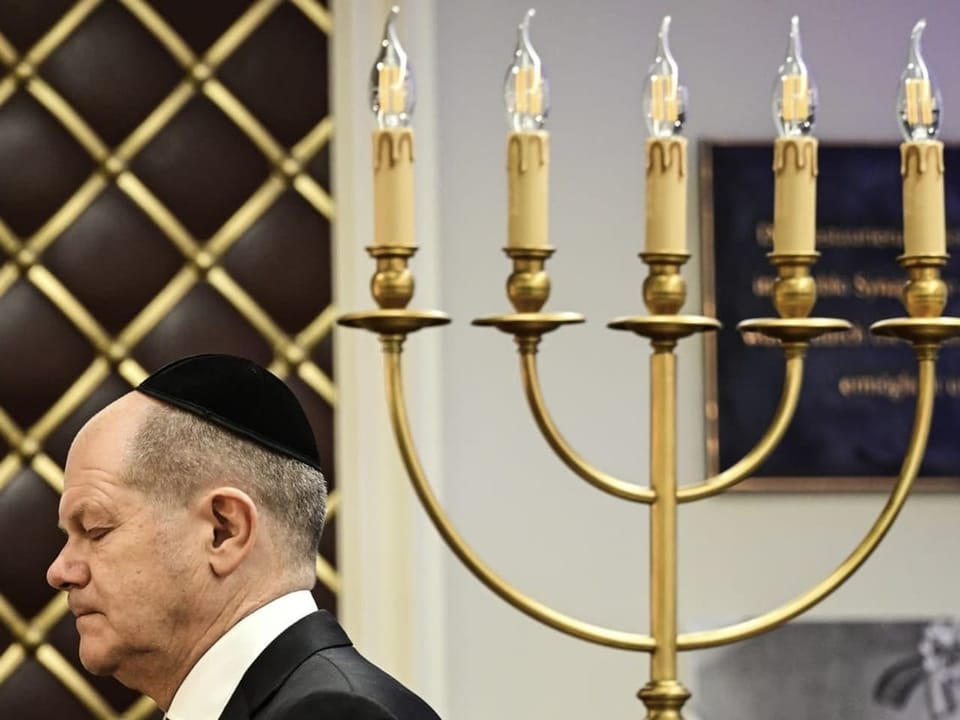 He walks past a candlestick towards a podium.  He wears a yarmulke.