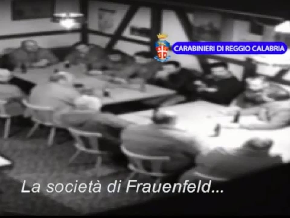 A dozen men sit at a table.  Screenshot of surveillance video.