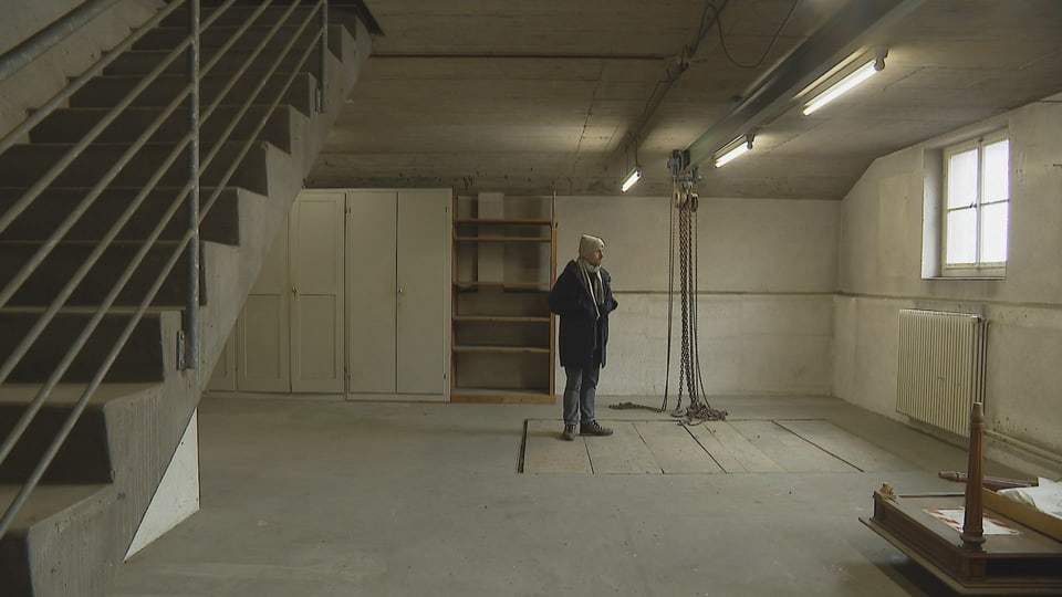 A man wearing a cap, scarf and coat stands in an empty, industrial-looking room.  A staircase leads upstairs.