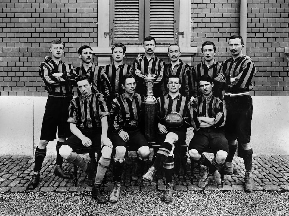 Black and white image with eleven men in striped jerseys gathered around a trophy.