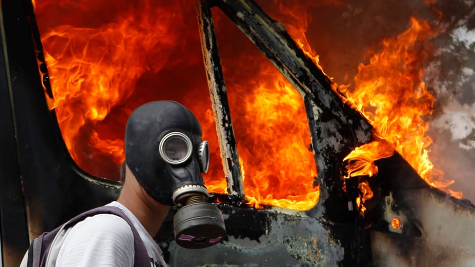 Symbolic image: person with gas mask in front of burning car.