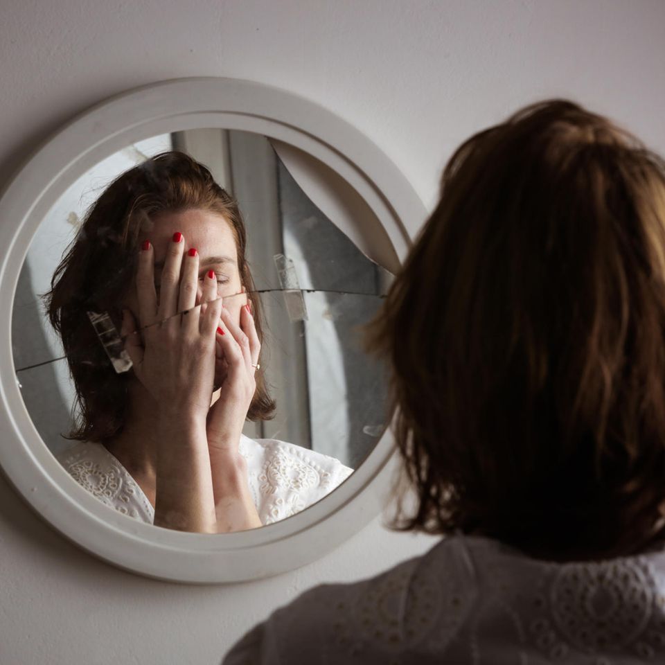 young woman stands in front of a broken mirror and holds her hands in front of her face