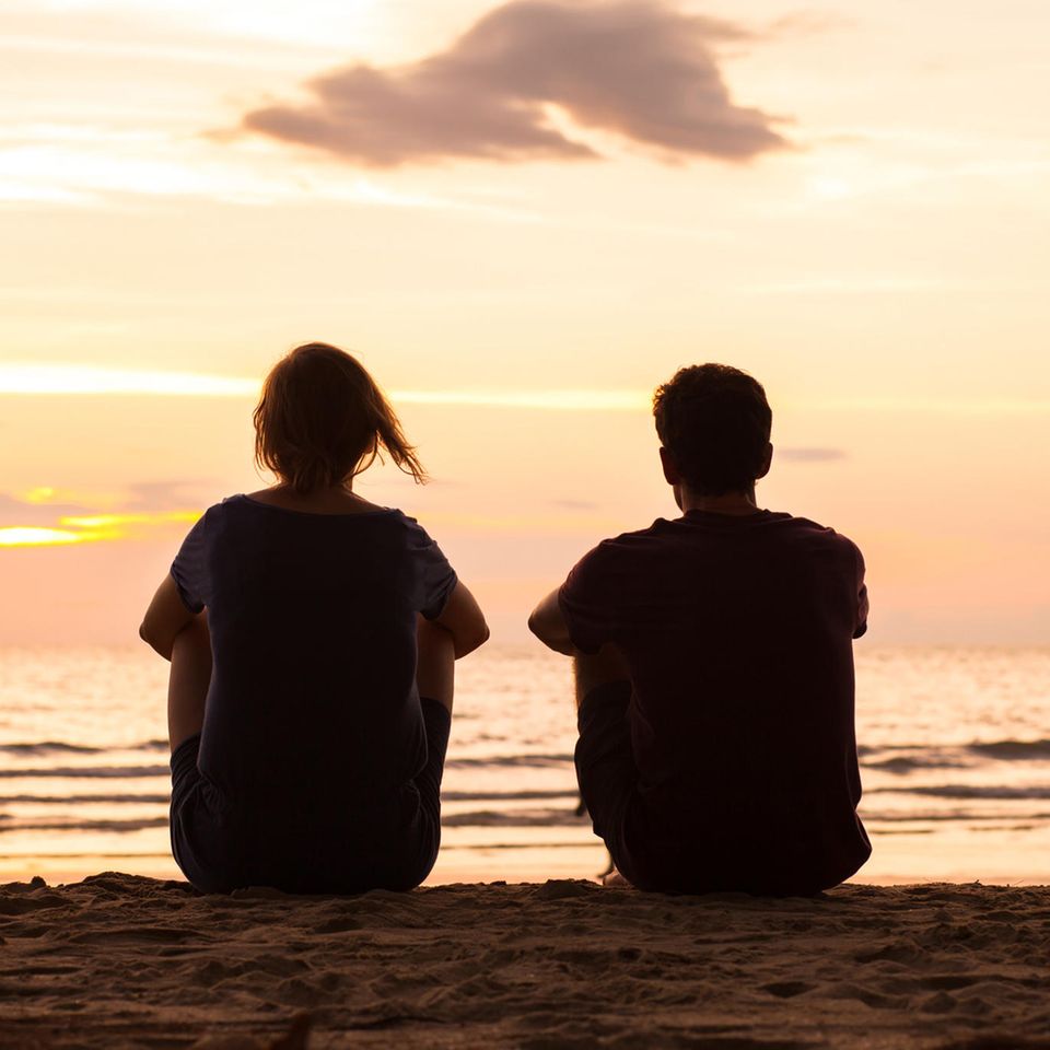 Two people sit on the beach in front of a sunset