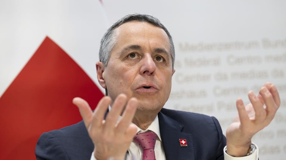 Ignazio Cassis, with short gray hair in a dark blue jacket, raises his hands in explanation.  Behind it is the Swiss flag.