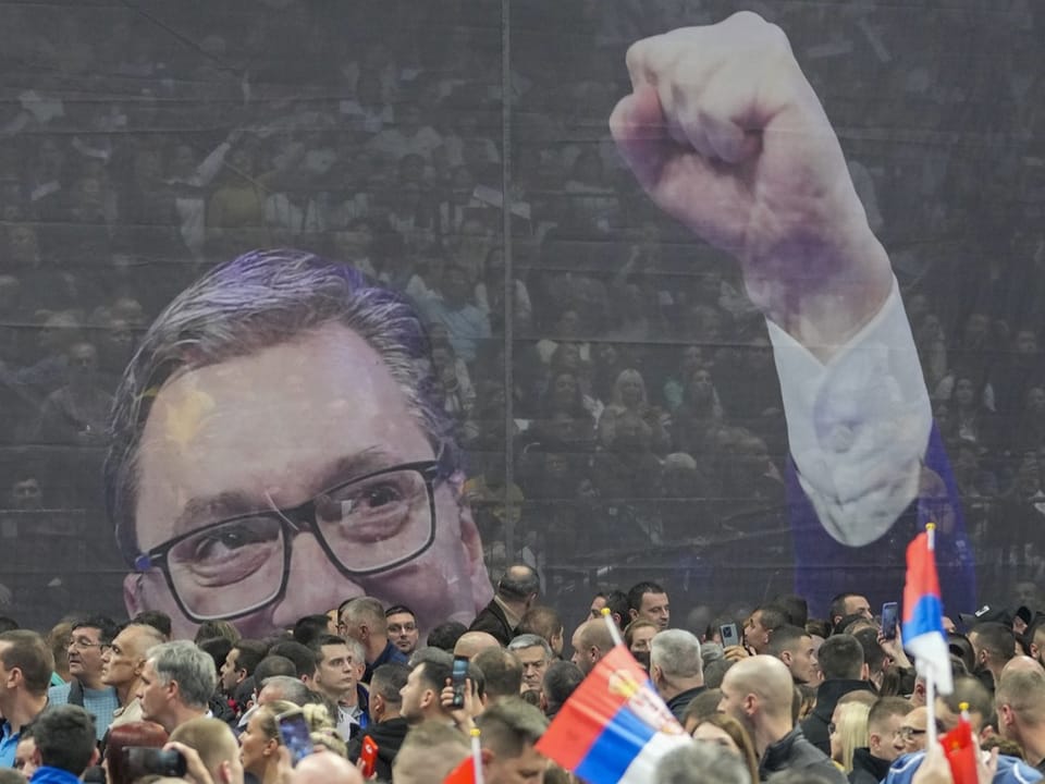 Crowd of people, some with Serbian flags, behind them a large projection of a man with glasses, raising his fist in the air