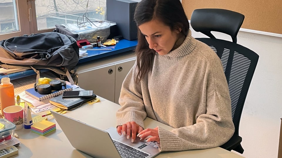 young woman with dark hair and a light gray sweater looking at a silver laptop while sitting at the desk.
