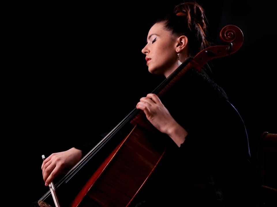 A woman with tied up brown hair plays the cello.  She has closed her eyes.