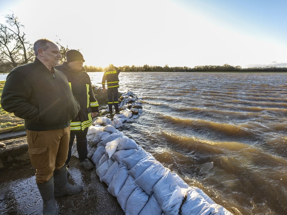 Men stand at a barrier with sandbags, behind a lake.