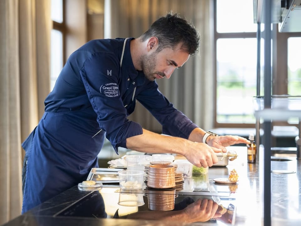 A man in a dark blue apron is concentrating on arranging a dish.